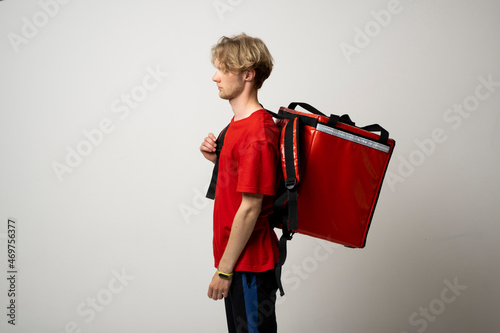 Young delivery employee man in red tshirt uniform with a thermal food bag backpack works as a courier in a food delivery service on a white background.