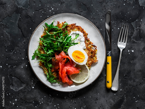 Large potato rosti with egg, arugula, salmon and Greek yogurt sauce on a dark background, top view. Delicious food