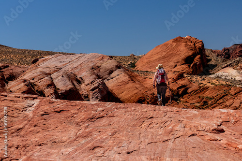 A female hiker walks across the Fire Wave of multi-colored rock layers that stand out amongst the red rocks and desert landscape in Valley of Fire State Park, Nevada 