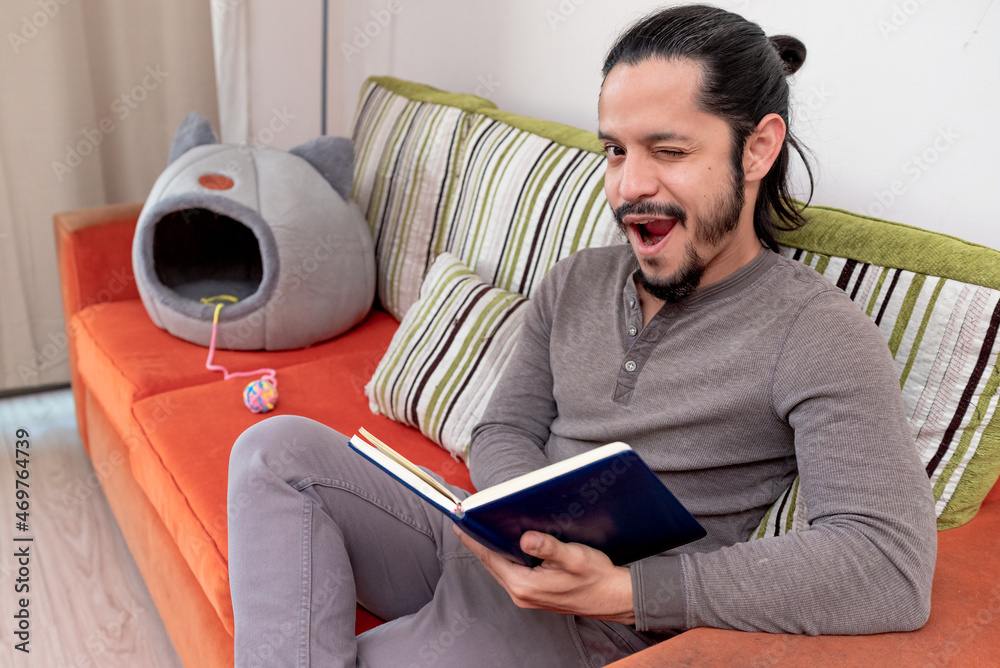 Young man reading a book at home sitting on the couch in the living room looking at camera Young man reading a book at home sitting on the couch in the living room looking at camera