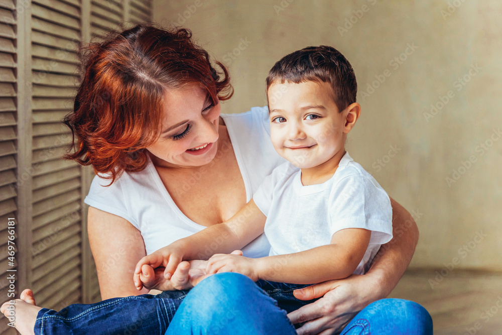 Young mother embracing her child. Woman and little boy relaxing at home near gray wall indoors. Happy family at home. Young mom playing with her son Young mother embracing her child. Woman and little boy relaxing at home near gray wall indoors. Happy family at home. Young mom playing with her son