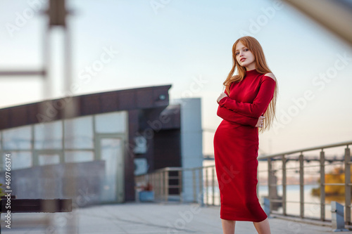 Portrait casual of a Red hair girl weared in a red dress, looking straight, arms folded, stays against blue sky outdoor. model attractive, long straight hair, fashion outfit. Blurred details