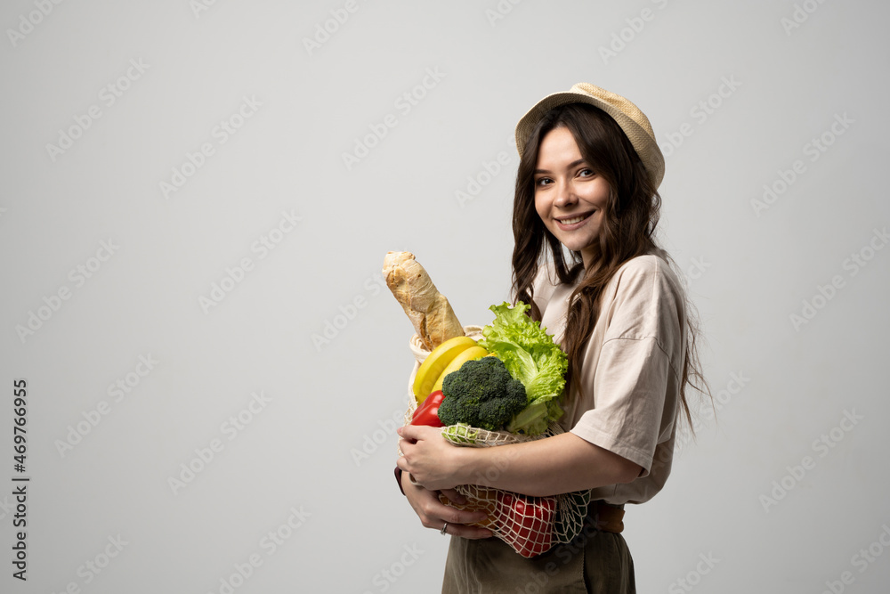 Fototapeta premium Portrait of happy smiling young woman in beige oversize t-shirt holding reusable string bag with groceries over white background. Sustainability, eco living and people concept.