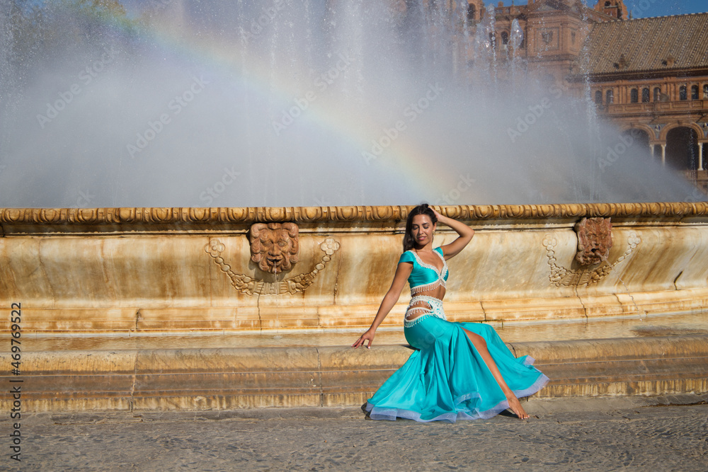 young and beautiful belly dancer sitting on the edge of a fountain in a large square. She is barefoot and her hair is loose. She is wearing a blue dress. Fashion and beauty concept young and beautiful belly dancer sitting on the edge of a fountain in a large square. She is barefoot and her hair is loose. She is wearing a blue dress. Fashion and beauty concept