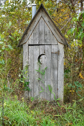 Rural wooden outhouse surrounded by trees, leaves and vines.     