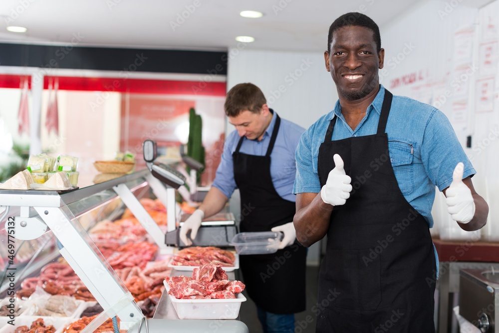 Happy African American seller of butcher store standing behind counter, giving thumbs up
