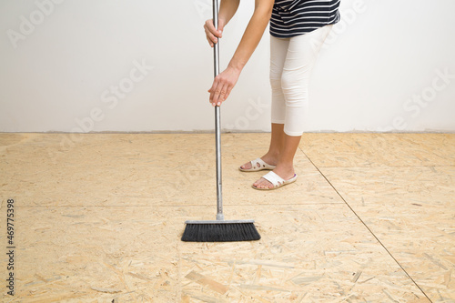 Young adult woman using broom and sweeping sawdust from osb board floor. Cleaning garbage after repair work of home. Front view. photo