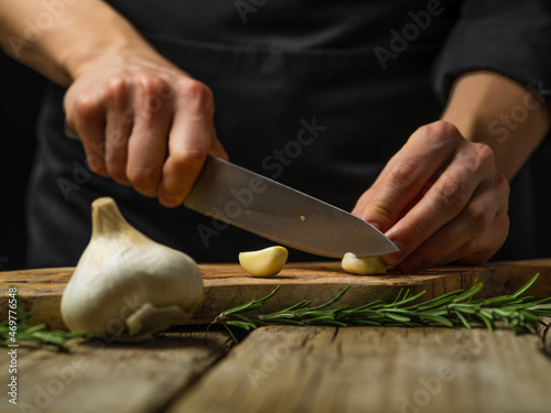 Close-up. The chef cuts the garlic on a cutting board. a sprig of rosemary lies nearby. Rough wooden table. Country style. Concept - cooking salad, sauce, soup. Restaurant, cafe, hotel.