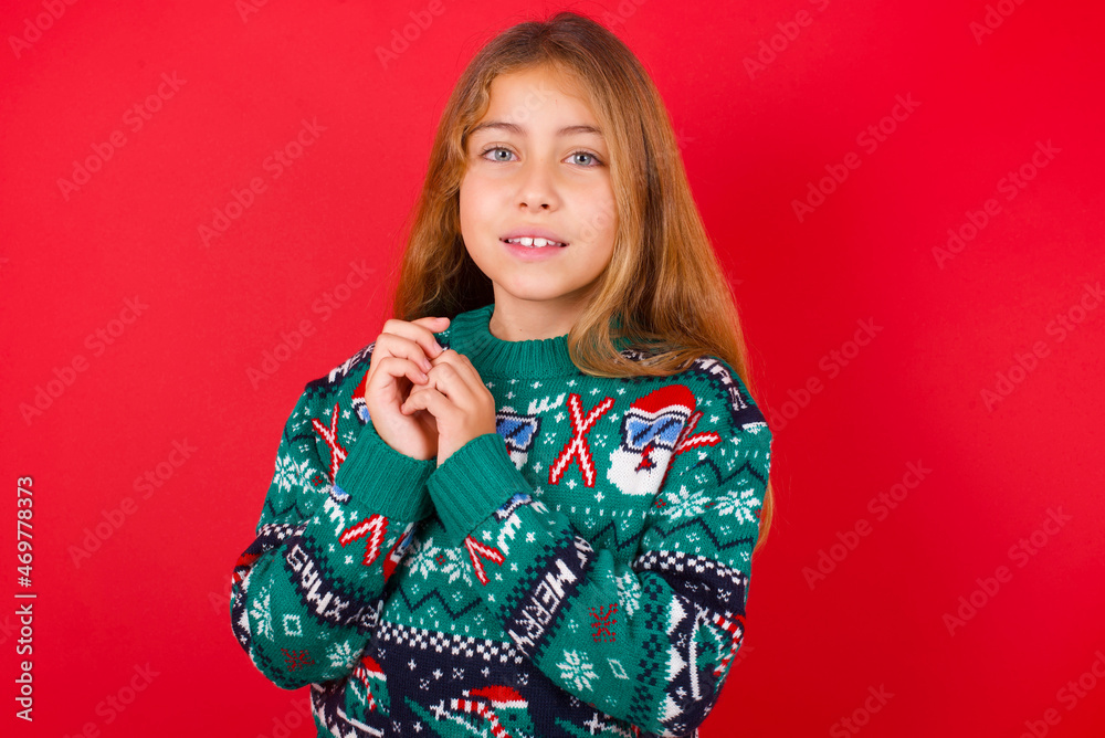 Dreamy charming little kid girl wearing knitted sweater christmas over red background with pleasant expression, keeps hands crossed near face, excited about something pleasant. Dreamy charming little kid girl wearing knitted sweater christmas over red background with pleasant expression, keeps hands crossed near face, excited about something pleasant.
