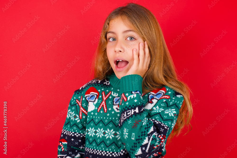 Shocked, astonished little kid girl wearing knitted sweater christmas over red background looking surprised in full disbelief wide open mouth with hand near face. Positive emotion Shocked, astonished little kid girl wearing knitted sweater christmas over red background looking surprised in full disbelief wide open mouth with hand near face. Positive emotion