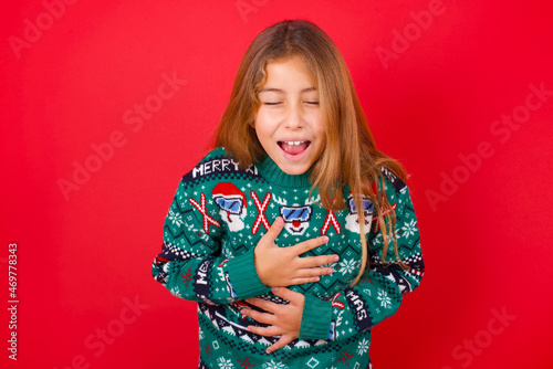 little kid girl wearing knitted sweater christmas over red background keeps hands crossed, laughs out loud at good joke, wears casual clothes.