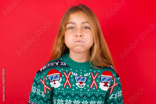 little kid girl wearing knitted sweater christmas over red background puffing cheeks with funny face. Mouth inflated with air, crazy expression.