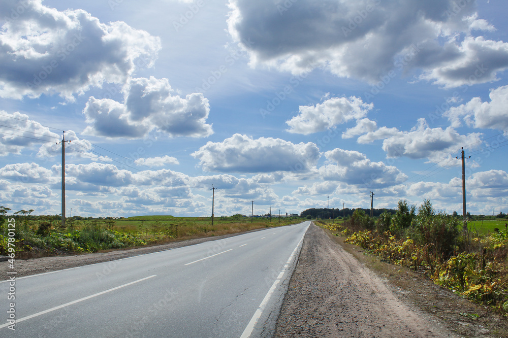 Empty highway under blue sky with clouds on sunny day, poles with electric wires and green grass on the sides