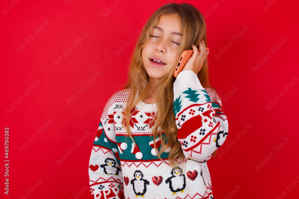 Funny little kid girl wearing knitted sweater christmas over red background laughs happily, has phone conversation, being amused by friend, closes eyes. Funny little kid girl wearing knitted sweater christmas over red background laughs happily, has phone conversation, being amused by friend, closes eyes.