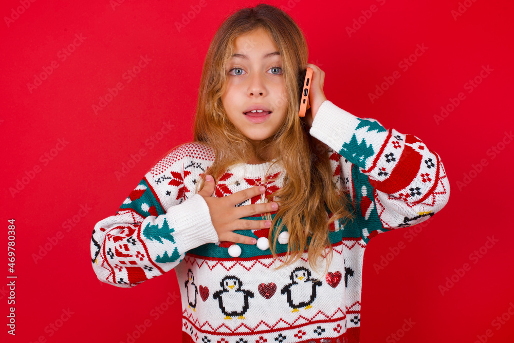 Smiling little kid girl wearing knitted sweater christmas over red background talks via cellphone, enjoys pleasant great conversation. People, technology, communication concept Smiling little kid girl wearing knitted sweater christmas over red background talks via cellphone, enjoys pleasant great conversation. People, technology, communication concept