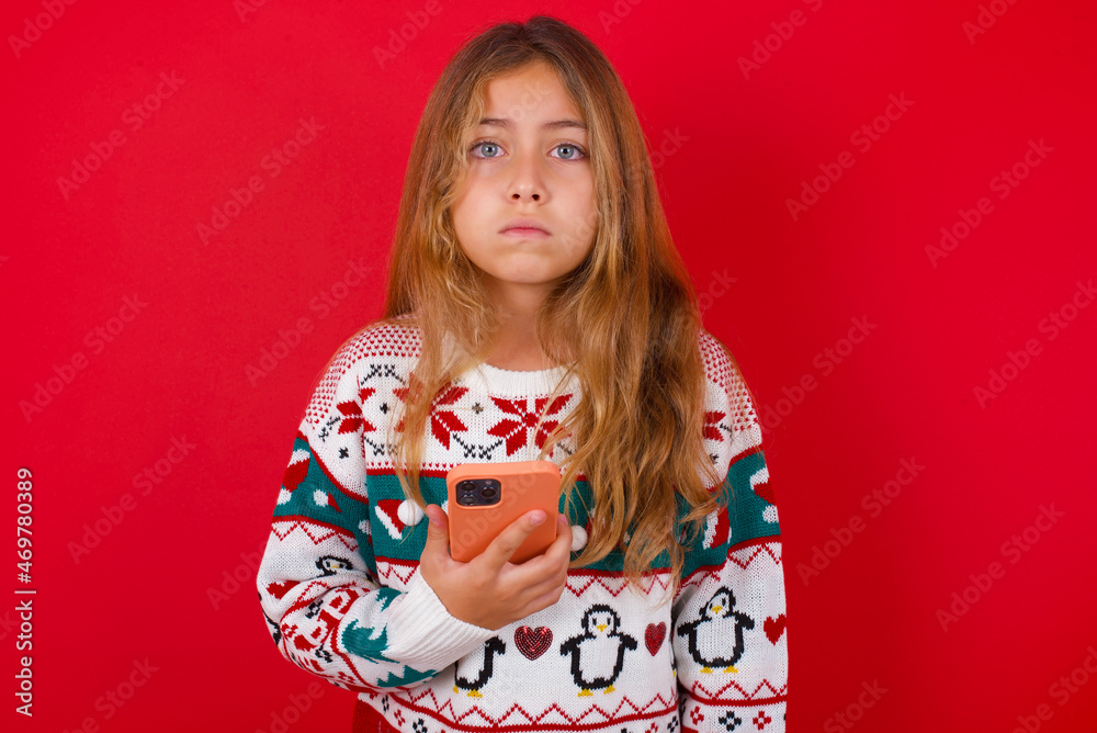 Upset dissatisfied little kid girl wearing knitted sweater christmas over red background uses mobile software application and surrfs information in internet, holds modern mobile hand