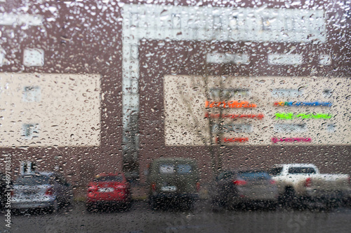 View through glass covered with raindrops to the building wall and parking lot with cars