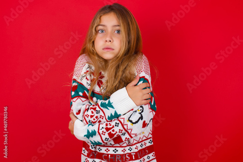 little kid girl wearing knitted sweater christmas over red background shaking and freezing for winter cold with sad and shock expression on face.