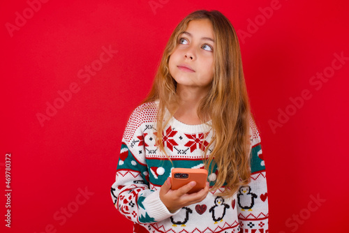 little kid girl wearing knitted sweater christmas over red background holds mobile phone uses high speed internet and social networks has online communication. Modern technologies concept