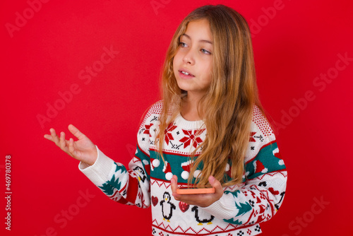 Happy pleased little kid girl wearing knitted sweater christmas over red background raises palm and holds cellphone uses high speed internet for text messaging or video calls