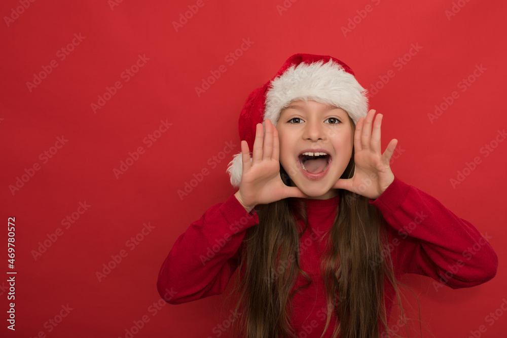 Little girl with long hair in red Christmas sweater and Santa hat is happy to shout. Shout. An emotional child stands with an open mouth and shouts on a red background. Human emotions, facial Little girl with long hair in red Christmas sweater and Santa hat is happy to shout. Shout. An emotional child stands with an open mouth and shouts on a red background. Human emotions, facial
