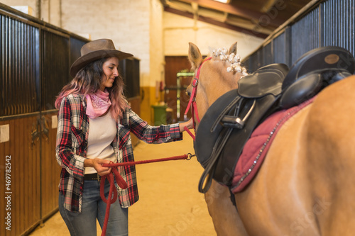Cowgirl woman petting a horse in a stable, wearing southern usa hats, pink plaid shirt and jeans