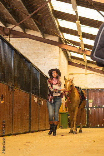 Caucasian cowgirl woman walking a horse in a stable, southern usa hats, pink plaid shirt and jeans