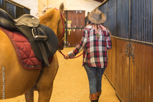 Unrecognizable cowgirl woman strolling with a horse in a stable, southern usa hats, pink plaid shirt and jeans