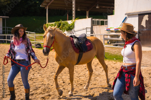 Two smiling cowgirl women pulling a horse out of a stable, in south american outfits