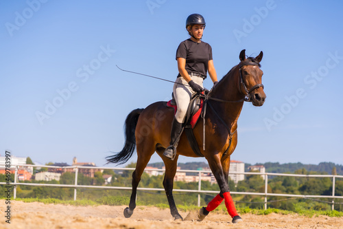 Young Caucasian blonde girl riding on a horse with a brown horse, dressed in black rider with safety cap © unai