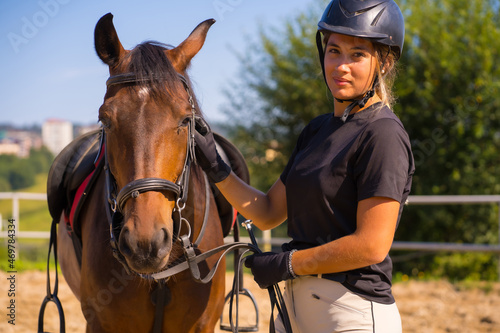 Posing of a Caucasian blonde girl on a horse caressing and pampering a brown horse, dressed in black rider with safety cap © unai