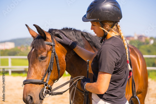 Posing of a Caucasian blonde girl on a horse caressing and pampering a brown horse, dressed in black rider with safety cap