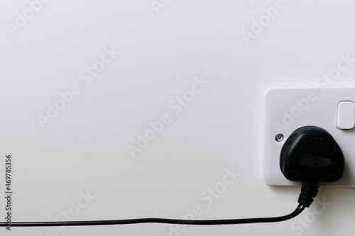 A black electric plug with cable in a white socket on a white wall. Copy space image. Electricity at home with 220 volt. Minimal concept. The socket is turned off because of the high energy prices.