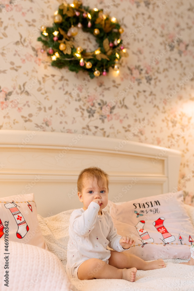 a child in white clothes sits on the bed and licks her hand. A Christmas wreath hangs over the bed, and there are Christmas pillows next to the child. a child in white clothes sits on the bed and licks her hand. A Christmas wreath hangs over the bed, and there are Christmas pillows next to the child.
