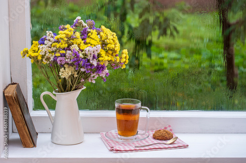 Old book, colorful bouquet of flowers and white cup of tea on background of window with raindrops at home at summer day