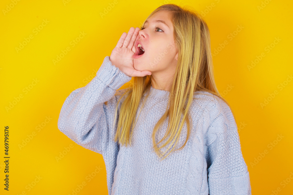 caucasian kid girl wearing blue knitted sweater over yellow background shouting and screaming loud to side with hand on mouth. Communication concept. caucasian kid girl wearing blue knitted sweater over yellow background shouting and screaming loud to side with hand on mouth. Communication concept.