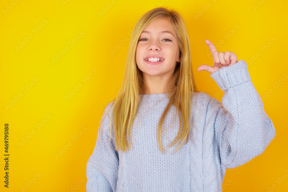 caucasian kid girl wearing blue knitted sweater over yellow background smiling and confident gesturing with hand doing small size sign with fingers looking and the camera. Measure concept caucasian kid girl wearing blue knitted sweater over yellow background smiling and confident gesturing with hand doing small size sign with fingers looking and the camera. Measure concept
