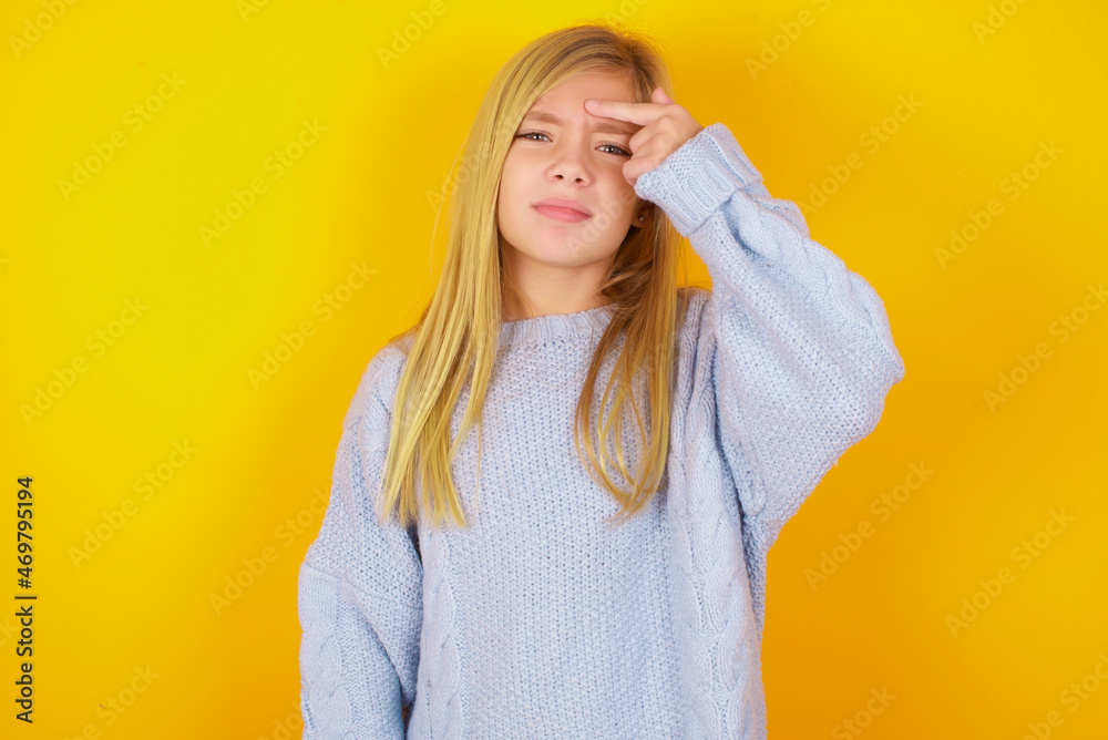 caucasian kid girl wearing blue knitted sweater over yellow background pointing unhappy at pimple on forehead, blackhead infection. Skincare concept. caucasian kid girl wearing blue knitted sweater over yellow background pointing unhappy at pimple on forehead, blackhead infection. Skincare concept.