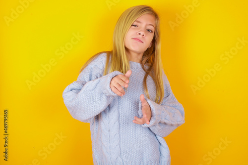Tablou pe pânză Joyful caucasian kid girl wearing blue knitted sweater over yellow background wink and points index fingers at camera, chooses someone, makes finger gun pistol