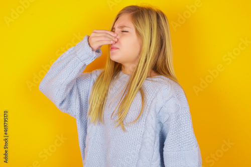 Fotografija caucasian kid girl wearing blue knitted sweater over yellow background  smelling something stinky and disgusting, intolerable smell, holding breath with fingers on nose