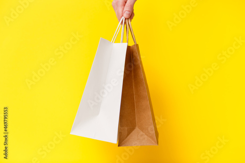 Female hand holding two shopping bags isolated on yellow background. White and brown craft blank paper bags in hand. Black friday sale, discount, shopping and ecology concept.