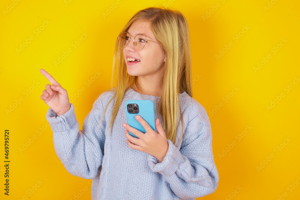 Wow!! excited caucasian kid girl wearing blue knitted sweater over yellow background showing mobile phone with open hand gesture Wow!! excited caucasian kid girl wearing blue knitted sweater over yellow background showing mobile phone with open hand gesture