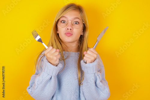 hungry caucasian kid girl wearing blue knitted sweater over yellow background  holding in hand fork knife want tasty yummy pizza pie