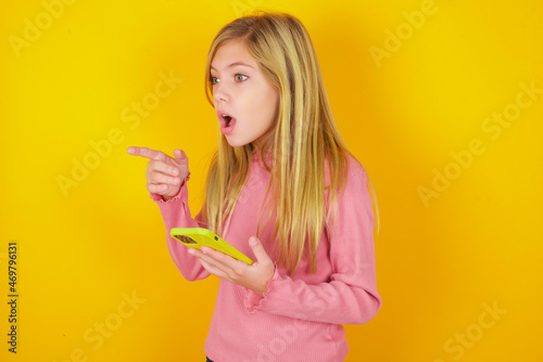 Stunned caucasian little kid girl wearing long sleeve shirt over yellow background points sideways right copy space  recommends product  sees astonishing thing