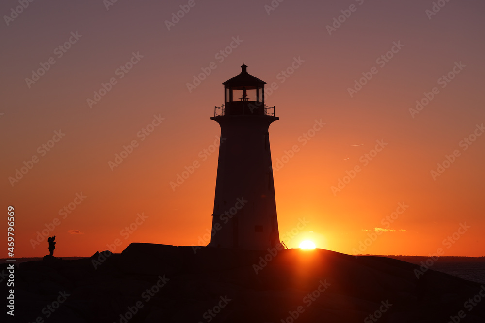 Silhouette shot of a lady taking a   picture of a lighthouse on a beautiful sunset full of red and orange sky. Peggy's Cove Lighthouse, Halifax, Nova Scotia, Canada
