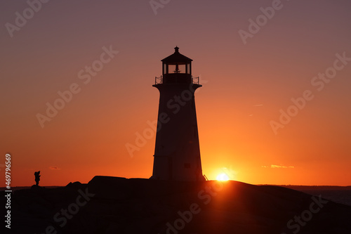 Silhouette shot of a lady taking a picture of a lighthouse on a beautiful sunset full of red and orange sky. Peggy's Cove Lighthouse, Halifax, Nova Scotia, Canada