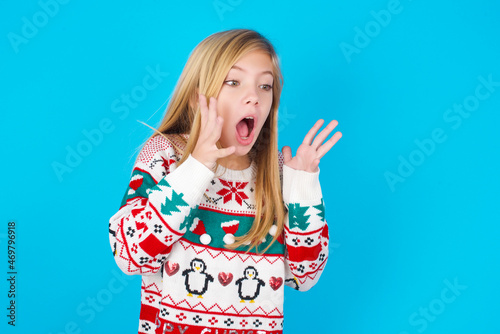 caucasian little kid girl wearing knitted sweater christmas over blue background with shocked facial expression, holding hands near face, screaming and looking sideways at something amazing.