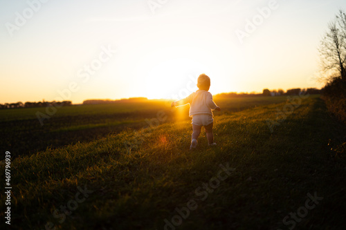 Golden hour photo of carefree toddler running off into the sunset in the green grassy meadow. Autumn, silhouette, sunset. © Jim
