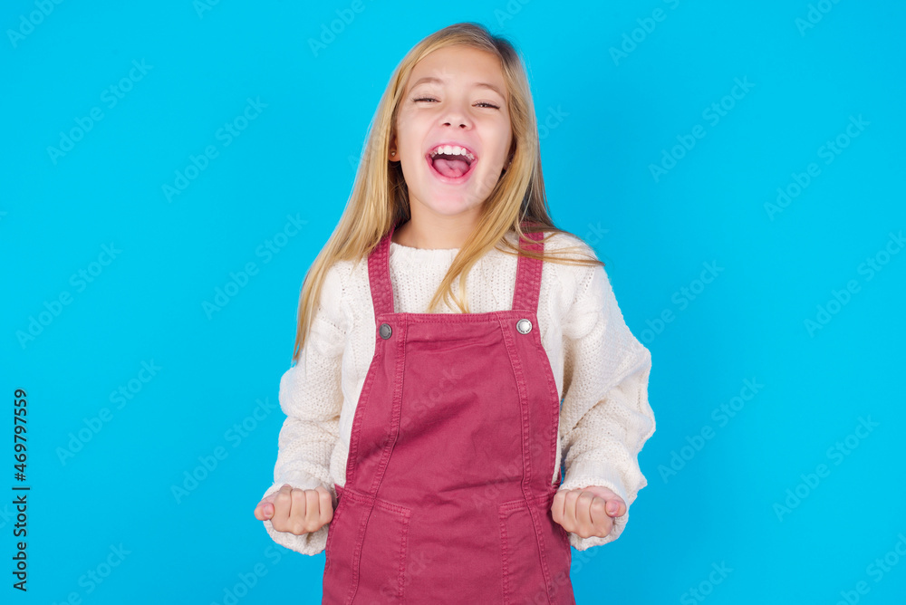 Fototapeta premium caucasian little kid girl wearing jumpsuit over blue background raising fists up screaming with joy being happy to achieve goals.