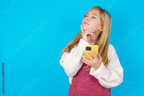 Thinking dreaming caucasian little kid girl wearing jumpsuit over blue background using mobile phone and holding hand on face. Taking decisions and social media concept.